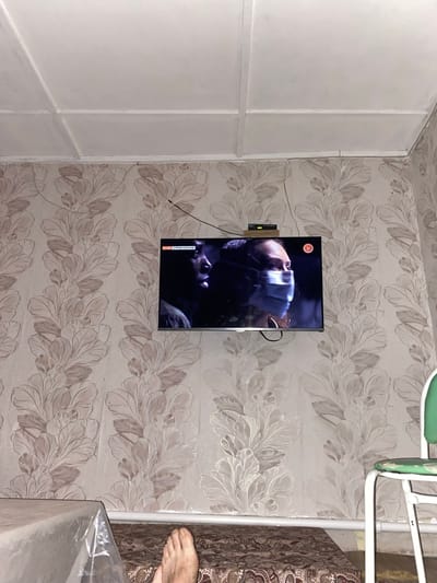 The image shows a television mounted on a wall with floral wallpaper. The television screen displays a man wearing a mask, with a slight blur suggesting motion. To the left of the TV, there's a black cable connected to a small grey device. A thin black wire also extends from the TV upwards and to the left. On the wall, partially visible, is the corner of a white, partitioned structure, possibly a closet or cabinet. To the right of the TV, a corner of a bed with patterned bedding is visible, and a human foot is propped up on it. The lighting appears to be indoor. The wallpaper has a repeating pattern of large, light-colored leaves against a slightly darker background. On the TV screen, in the lower left corner, there is text that reads "СЛЕЗЫ СОЛНЦА". The image is oriented vertically.