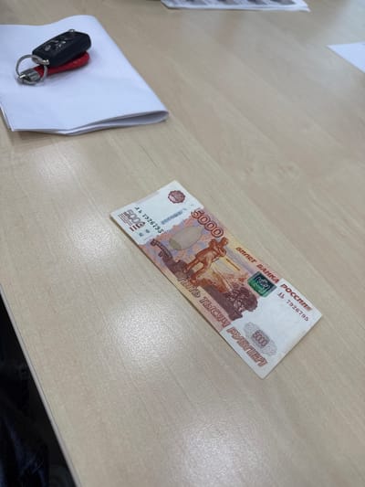 A 5000 ruble note is lying on a light-colored wooden desk. A car key with a red tag is on top of a stack of white paper to the left of the note. The image does not provide any information about the city of Емельяново, Russia.