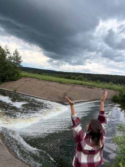 The image captures a scene at a waterfall in Nizhnii Vishur, Russia. A woman in a red and white plaid shirt stands at the edge of a concrete structure, presumably a dam, reaching her arms out towards the cascading water. The waterfall is the primary focal point. Behind the dam is a grassy field and a line of trees, suggestive of a forest. Above, a cloudy sky dominates the view. The overall perspective suggests the photo was taken at an angle.