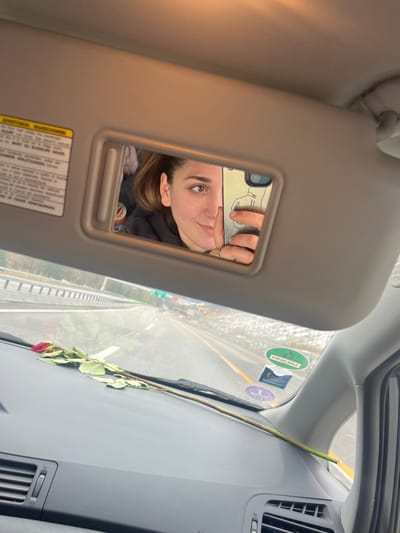 The image depicts the interior of a vehicle on a highway, observed from the passenger side. A lowered passenger sun visor reflects a female individual in its integrated mirror, actively taking a selfie with a smartphone held in her right hand, partially obscuring her face. She has brown hair, brown eyes, and a slight smile, and two rings are visible on her right ring finger. The smartphone case is light-colored and features a line-art illustration. A multi-language warning label is present on the sun visor. The vehicle's black dashboard, equipped with air vents, holds a single red rose with a long green stem and leaves, positioned horizontally with the bloom towards the left. Through the windshield, a multi-lane highway is visible, appearing wet and bordered by guardrails. Patches of snow or ice are present along the roadside. Two circular stickers, one green with a crescent moon and star and one purple, are affixed to the lower right corner of the windshield. The scene is located on a highway near Airolo, Switzerland.