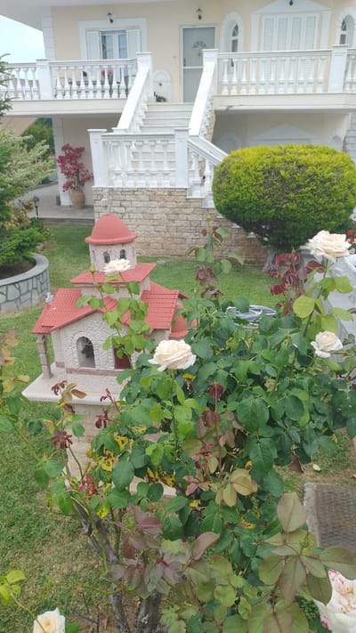 In the foreground of the image, a miniature replica of a church with a red tiled roof and a dome is visible. The church is adorned with a small cross on its roof. Several light-colored roses are blooming in front of and around the church replica. The background features a multi-story building with white railings and stone accents. To the right, a large, rounded green bush is present, and to the left, a potted plant with red leaves sits near a stone wall. The scene is set in a garden with green grass. The location is identified as Νέα Κερασιά, Greece. There are no people or animals visible in the image.