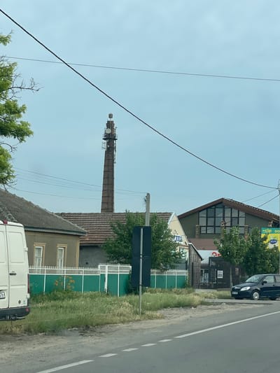 In Congaz, Moldova, a scene unfolds beneath a cloudy sky, featuring a towering brick chimney converted into a telecommunications tower. The tower, adorned with antennas and various equipment, stands prominently against the overcast heavens. At the base of the chimney, residential buildings with tiled roofs are visible, some featuring traditional architectural elements. A white van is parked on the left side of the image, and a dark blue car is positioned on the right, near the edge of a paved road with white markings. A green fence separates the buildings from the roadside, with overgrown grass and vegetation growing in front of it. The overall impression is one of a rural or semi-urban environment where old industrial structures have been repurposed for modern technological use.