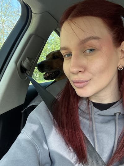 A woman with red hair is in a car, wearing a grey hooded sweatshirt and a seatbelt. A dog is partially visible behind her, peering into the car from outside. The car's interior is visible, including the door and window frame. Outside the car, trees and foliage are visible. The location of the photo is not determinable from the image alone.