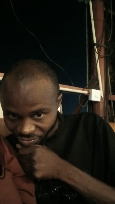A man with a shaved head and dark skin is looking at the camera. He is wearing a black t-shirt. His left hand is raised near his mouth. In the background, there's a dark blue wall and a ceiling with exposed wiring and light fixtures. The lighting is dim and the image appears to be taken indoors. The photo is rotated.
