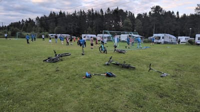 In Östersund, Sweden, on a grassy field with scattered dirt patches, a group of individuals, predominantly young people, are engaged in a casual soccer game near a goal net. Several bicycles and scooters are lying on the ground, some appearing to have been dropped or fallen over. In the background, a line of recreational vehicles, including caravans and motorhomes, are parked, suggesting a campground or similar facility. Behind the vehicles, a dense forest of coniferous trees forms the horizon under an overcast sky. The overall scene depicts a recreational area where outdoor activities are taking place.