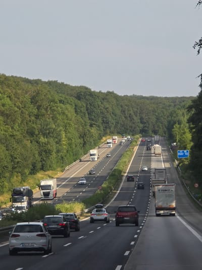 The image depicts a highway scene in the vicinity of Bremthal, Germany. A multi-lane highway is visible, flanked by dense green trees on either side. The highway is busy with various vehicles, including cars and trucks, traveling in both directions. Some of the vehicles are passing each other, while others are maintaining their lanes. There is a speed limit sign visible on the right side of the highway, indicating a restriction of 100 km/h. A blue road sign on the right indicates directions to Würzburg and Frankfurt. The sky is overcast, and the overall lighting suggests it is daytime. The terrain appears to be somewhat hilly, with the trees covering the slopes.