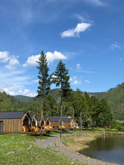 The image depicts a row of modern wooden cabins with black pitched roofs situated on a wooden deck beside a tranquil body of water. The cabins feature large, angled windows offering views of the surrounding natural landscape. Several Adirondack chairs and small grills are placed on the decks, suggesting a recreational setting. Tall evergreen trees flank the cabins, and the background is dominated by lush, green forested hills under a bright blue sky with scattered white clouds. The scene appears to be a resort or glamping site located in the scenic environment of Bol'shoe Goloustnoe, Russia, likely on the shore of Lake Baikal or a nearby body of water.