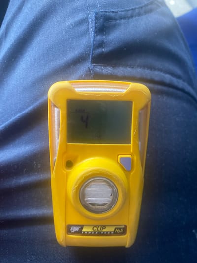 A yellow BW CLIP H₂S gas detector is positioned on a person's dark blue fabric clothing. The detector's screen displays "MM" and the number "4". The device has a circular sensor at the bottom and a purple button on the right side. The clothing appears to be a uniform, likely workwear, given the presence of the gas detector. The background consists of the textured fabric of the clothing. The scene is indoors or in a controlled environment, with no visible windows or outdoor elements. The image does not provide information about the specific location within Қосшағыл ауылдық округі, Kazakhstan.