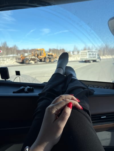 The image displays a scene viewed from the interior of a vehicle in Staroe Mikhailovskoe, Russia, looking forward through the windshield on a clear, sunny day.
In the foreground, a person's legs, clad in dark trousers and light gray socks, are crossed at the ankles and rest on the dark-colored dashboard. Below the legs, a right hand with bright red polished fingernails and a silver-toned ring on the ring finger is visible, resting on the person's lap. The vehicle's dashboard features a dark, textured surface, air vents, and a brown trim. Small black electronic devices or mounts are affixed to the dashboard near the windshield. Partially visible Cyrillic text, "ОТОЙ," is present on a label near the bottom of the windshield.
Outside the vehicle, a paved road is visible, flanked by residual snow on the shoulders. A large yellow road grader or construction vehicle is positioned on the right side of the road, appearing stationary. Further down the road, a white box truck, featuring the Cyrillic text "АВТОПЕРЕВОЗКИ" (Auto Transport) and the numbers "64-89-47" on its side, is also visible. Bare deciduous trees line the distant horizon under a bright blue sky with faint cirrus clouds. A red and white road sign is partially visible on the far left.
