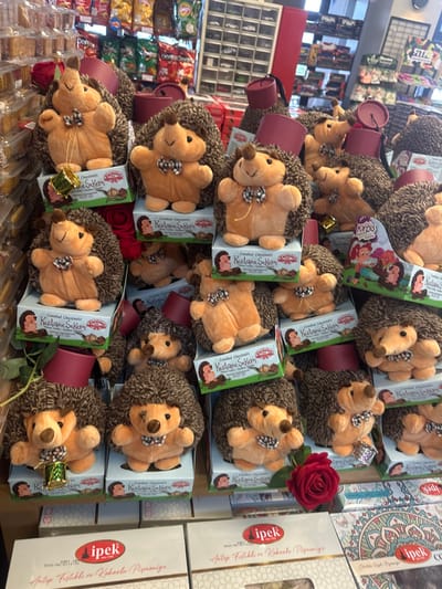 This image depicts a retail product display located within a commercial establishment in Kemalpaşa, Turkey. The central focus is a multi-tiered arrangement of approximately three dozen plush hedgehog toys. Each hedgehog, characterized by beige fur and dark brown shaggy "spines," is adorned with a black-and-white checkered bow tie. Many of these toys are also accessorized with small, red fez-like hats or miniature golden drums. Each plush hedgehog rests atop a light blue/green cardboard box labeled "Candied Chestnuts KESTANE ŞEKERİ," with "Sardesli" and "Kemalpaşa" branding visible. Additional "Kirpis" branded boxes featuring cartoon hedgehogs are also present. Artificial red roses are strategically placed throughout the display as decorative elements. In the lower foreground, larger white boxes branded "ipek" are visible, with text indicating "Antep Fıstıklı ve Kakaolu Pişmaniye." The background reveals shelves stocked with various packaged snack foods, including branded potato chips, and other retail shelving units. No individuals are present, and no actions are occurring within the static commercial display.