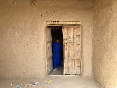 The image shows the interior of a mud-brick building in Saranan, Pakistan, located at 30.532 latitude and 66.909 longitude. A young boy, wearing a blue shirt and pants, stands in the doorway of the building, looking out at the viewer. The doorway is comprised of two wooden doors that are open. There are numerous pieces of trash scattered on the dirt floor in front of the doorway. The interior wall of the building is made of mud brick and has white writing scrawled on it. There are also some decorative carvings above the doorway. The boy appears to be smiling as he looks directly at the viewer. The image was likely taken from inside the building.  The overall impression of the scene is one of poverty and simplicity.