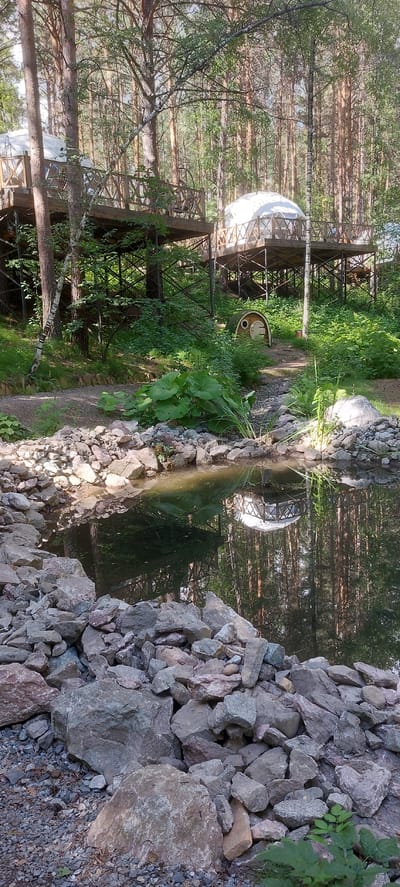 The image depicts a tranquil natural setting with man-made structures nestled within a forest. The primary subjects include a geodesic dome-like structure, possibly a dwelling or a unique architectural feature, situated amongst tall trees. Beside it, a small, rounded wooden building with a door is partially visible, peeking out from lush greenery.
The scene is characterized by a reflective body of water, likely a pond or a small lake, with rocks lining its edge. The water mirrors the surrounding trees and the man-made structures, creating a sense of depth and harmony. The forest is dense with a variety of trees, their trunks and branches creating a textured backdrop.
There is no discernible activity or event taking place; the image captures a still moment, suggesting a peaceful, possibly secluded, location. The time of day appears to be daytime, with natural sunlight filtering through the trees, casting dappled shadows and highlighting the green foliage. The weather seems pleasant, conducive to outdoor exploration or relaxation.
Notable details include the rustic wooden structures, the natural rock formations around the water, and the reflective quality of the water surface. The overall atmosphere is serene and natural, with a touch of unique design elements introduced into the landscape. There is no visible text in the image. The location context provided places this scene in Berdsk, Russia, suggesting a possible recreational or residential area within a forested environment.