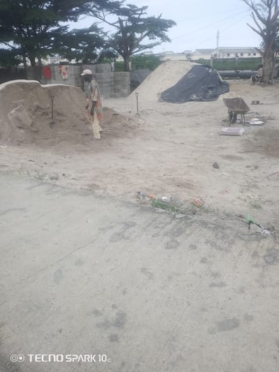 In Adesanya, Nigeria, a construction site is depicted under an overcast sky. The scene is dominated by piles of sand, a common material in construction. A man, dressed in a patterned shirt and light-colored trousers, stands near a large sand dune, seemingly involved in the construction activities. His attire suggests a practical choice for the environment.

Other objects present include stacks of concrete blocks, indicating the building materials readily available on site. A wheelbarrow, a shovel, and a large black tarp covering some materials are also visible, all essential tools and elements of a construction zone. In the background, trees provide a natural backdrop, with some residential buildings and power lines suggesting the site is in a populated area.

The overall impression is one of ongoing work, with the scattered debris and materials hinting at the dynamic nature of a construction project. The time of day appears to be daytime, based on the ambient light, though the cloudy weather obscures direct sunlight. The scene is characteristic of many developing areas where construction is actively underway. No specific emotional cues are evident from the subjects. The text "TECNO SPARK 10c" is visible at the bottom left corner of the image.