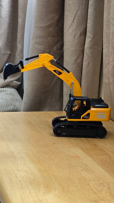 A bright yellow toy excavator with black tracks sits on a light wooden surface, which appears to be a tabletop. The excavator is positioned with its boom and bucket extended, resting against a background of textured beige fabric, possibly upholstery or curtains. The lighting suggests it is daytime. The scene is static, with no active event taking place. A small orange mark is visible on the wooden surface near the excavator. There is text visible on the excavator's arm and body, featuring the word "GEAR". The context of Kilkenny, Ireland, is provided but not visually evident from the image itself.
