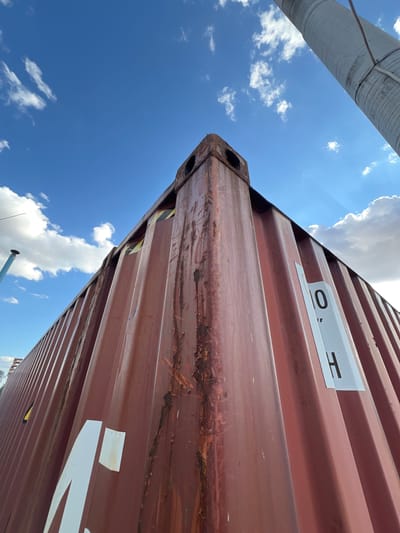 This image captures a close-up, low-angle view of a large, rusty, corrugated metal shipping container against a bright blue sky dotted with fluffy white clouds. The container is a deep maroon or brown color, showing signs of wear and tear with visible rust streaks and some peeling paint. A white label with black stenciled letters "O H" is affixed to the side of the container. Another larger white marking, partially visible, appears to be part of a word or logo.
The perspective is from below, looking upwards, emphasizing the massive scale of the container. Part of a large, ribbed, light-colored cylindrical structure, possibly a pipe or a silo, extends into the frame from the top left, also angled against the sky. The bright sunlight suggests it is daytime, likely mid-morning or afternoon, and the clear sky with scattered clouds indicates fair weather.
The scene appears to be an outdoor industrial or port area, given the presence of the shipping container. There are no people visible. The overall impression is one of industrial infrastructure under a clear sky. The location context provided is Újezd u Brna, Czechia, which might suggest a logistics hub or a similar facility in that region.