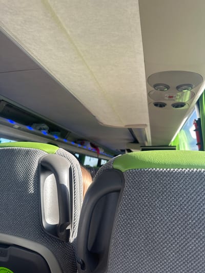 The image displays the interior of a public transport vehicle, likely a train or bus, captured from a passenger's perspective in Hattencourt, France. The scene is brightly lit, suggesting daytime.

In the foreground, the back of a modern passenger seat occupies the right side of the frame. This seat is upholstered in a textured, dark gray fabric, accented by a vibrant lime green trim along its contours. Two dark gray plastic handles are integrated into the seatback; one is positioned near the top, and another lower down. The upper part of the seat partially obscures the head of a person with light-colored hair, seated directly in front of the camera.

To the left of the seat, a long, light-colored (off-white or light gray) overhead panel stretches across the vehicle's interior. This panel features several circular components, appearing to be air vents or individual reading lights, some with mesh covers. One cluster of these circular elements prominently displays a small red indicator light in its center. Below this main panel, a light-colored, possibly cream-colored, fabric window blind or shade is pulled down. Further along the side of the vehicle, underneath the blind, a narrow storage compartment or shelf is visible, illuminated by a series of small, distinct blue LED lights. Within this compartment, several indistinct items, possibly bags or clothing, are stowed.

A glimpse of blue sky and some green structures are visible outside, through what appears to be a window above the overhead panel, confirming the daytime setting. There is no discernible text visible in the image.