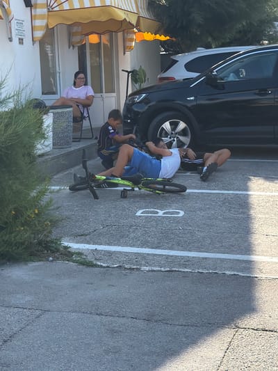 This media file shows two young boys and a woman in an outdoor setting that appears to be a parking lot or paved area in Općina Baška, Croatia. One boy, wearing a purple and black soccer jersey and blue shorts, is sitting on the ground next to a bicycle. The other boy, in a white t-shirt and blue shorts, is lying down with his legs up in the air, near a car. A woman is seated on a chair under a striped umbrella, looking towards the camera. The scene is brightly lit, suggesting it is daytime, and the presence of umbrellas might indicate warm weather. There are parking lines visible on the asphalt, and a car is parked to the left of the frame. A bicycle is positioned between the two boys. The background includes some greenery and what looks like a building with windows and doors. The overall mood seems relaxed and casual. There is a visible number "8" painted on the pavement.