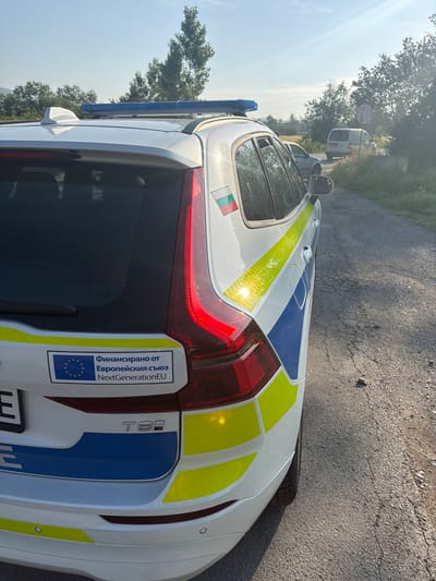 An overhead, tilted shot captures the rear and side of a white Volvo T8 estate or SUV, likely a law enforcement or official vehicle, parked on an asphalt road in Han Krum, Bulgaria. The car features distinctive blue and reflective yellow striped livery, common on police or service vehicles, along with a blue emergency light bar mounted on the roof. A small, three-colored emblem (green, white, red) with additional insignia is visible on the side near the rear window.

A prominent sticker on the rear window declares in Bulgarian: "Финансирано от Европейския съюз NextGenerationEU" (Funded by the European Union NextGenerationEU), accompanied by the European Union flag, indicating its funding source. The car also has a "T8 Recharge" badge, signifying its hybrid powertrain. Rear parking sensors are visible on the bumper.

The setting appears semi-rural or roadside, with lush green trees lining the road under a bright, sunny sky. Another white van and parts of a second vehicle are visible further down the road. The asphalt road surface shows some cracks. The bright sunlight suggests it is daytime, possibly morning or afternoon. No people are visible, and the car appears stationary.