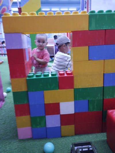 The image captures a brightly colored, large-scale building block structure in what appears to be an indoor play area. Two young children are visible within or near the structure. On the left, a child wearing a pink shirt is looking towards the camera. On the right, a child wearing a grey cap and a striped shirt is also visible. The building blocks are arranged in a playful, irregular fashion, forming an opening through which the children are seen. The floor is covered in green artificial turf, typical of indoor playgrounds. The background shows more colorful elements of the play area, including other structures and possibly seating. The overall scene is one of a child-friendly environment. The location is identified as Talas City, Kyrgyzstan.
