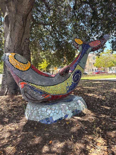 The image shows a colorful mosaic sculpture of a whale, positioned next to the trunk of a large tree. The sculpture is adorned with various shades of blue, green, yellow, red, and purple tiles, arranged in intricate patterns. The whale rests on a base also covered in mosaic tiles, some of which appear to have inscriptions. In the background, there are residential buildings and parked cars. The ground around the sculpture is covered with dirt, fallen leaves, and sparse vegetation. The overall scene suggests an outdoor public space, possibly a park or a street corner in a residential area. The presence of the tree and the buildings indicate an urban environment. The lighting suggests it is daytime, with sunlight filtering through the tree canopy. The sculpture appears to be a permanent installation, likely a piece of public art.