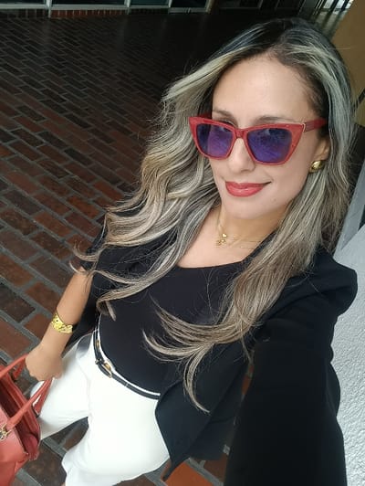 A woman poses in a selfie. She wears red sunglasses, a black top, and a black blazer. Her pants are white with a black and white belt. She has blonde and brown hair styled in waves, along with gold earrings, a gold necklace, and a gold bracelet. She is holding a red bag in her left hand. The background shows a brick floor and a white wall.