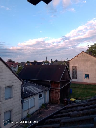 The image captures a suburban scene in Lohmar, Germany, likely during late afternoon or early evening due to the soft, warm lighting and the colors of the sky. The sky is a prominent feature, a pale blue with scattered, wispy clouds tinged with pink and orange, suggesting sunset or sunrise.

The scene is dominated by a collection of buildings and structures. In the foreground, a dark, tiled rooftop slopes downwards. To the left, the side of a light-colored, multi-story building is visible, with at least two windows. Next to it, a smaller, single-story building with a corrugated roof and closed shutters is seen. A dark wooden shed with a sloping roof and double doors stands in the middle ground. To the right, a larger, light-colored building with a pitched roof features a tall, rectangular window made of textured glass panes and a smaller square window below it that appears to have a framed picture or artwork.

Behind these structures, other rooftops and buildings are visible, interspersed with trees. A church spire, topped with a cross, is visible in the distance, hinting at the presence of a church or cathedral. Power lines stretch across the upper portion of the image, supported by a utility pole. At the base of the shed, several trash bins are lined up.

The overall mood is quiet and peaceful, reflecting the calm of a residential area at dusk. There are no people visible. The weather appears to be fair, with clear skies. The text "Shot on AWESOME A70" is visible in the bottom left corner of the image, indicating the device used to capture the photograph.