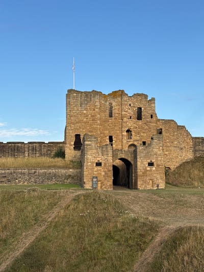 A ruined stone castle, located in North Tyneside, United Kingdom, stands under a clear blue sky. The structure is composed of light brown, cut stone blocks and displays significant architectural wear and decay, with a prominent central keep and connecting curtain walls. Multiple rectangular and arched openings are visible throughout the weathered facade, some appearing as empty window apertures and others as barred recesses. A large, dark arched gateway serves as the primary entrance, piercing the central lower portion of the structure. A white flagpole, topped with a partially furled or indistinct flag displaying red, white, and blue, consistent with the Union Jack, extends from the upper left battlements. The immediate foreground consists of uneven, dry grassy terrain, marked by two visible dirt paths ascending towards the castle's base. To the left of the main archway, a dark information sign is affixed to the stone wall. The scene is devoid of human or animal presence. The lighting is natural, suggesting daytime.