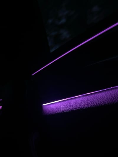 The image depicts a dark interior space, likely of a car, illuminated by purple lights. The texture of a material resembling leather is visible along the right side. The location is not discernible from the image.