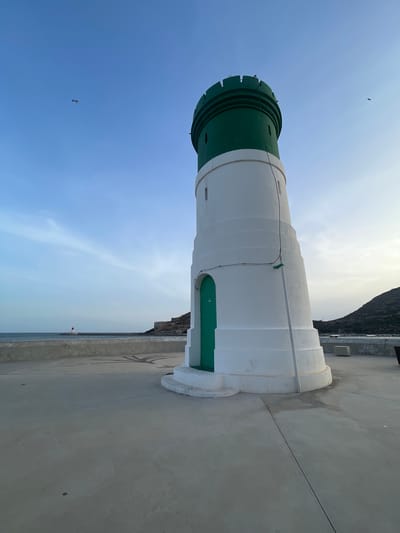 The image depicts a lighthouse with a white cylindrical body and a green top, situated on a concrete pier. The lighthouse has a green door at its base and is surrounded by a clear blue sky with wispy clouds. In the distance, the sea is visible, with a coastline and hills forming the background. The scene is devoid of any human presence. The location is not the city of Cartagena, Spain.