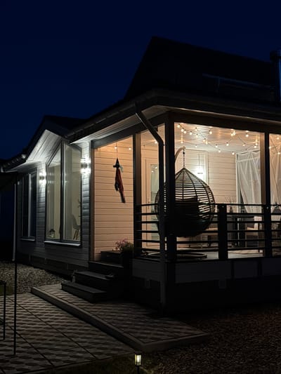 An exterior, low-angle shot depicts a house at dusk in Aleshkino DP, Russia. The scene is illuminated by string lights draped across the porch ceiling and two wall-mounted lights by the windows. A dark, woven egg-shaped hanging chair is suspended from the porch. A dark-colored roof overhangs the structure. The house features light-colored siding and large windows. A small, decorative item, possibly a witch's hat, hangs from the porch. A few steps lead up to the porch from a paved walkway. The ground surrounding the walkway is covered in small, dark pebbles. The sky is a deep, dark blue, indicative of twilight or night.