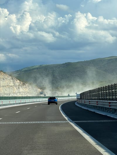 The image depicts a highway, likely a new construction or under development, curving through a mountainous landscape. A blue car is seen driving on the highway, facing away from the camera. The road is asphalt with clear lane markings and safety barriers on either side. The left side of the highway features a steep, rocky incline with visible layers of stone, suggesting excavation or a cut through the mountainside. The right side of the highway is bordered by a dark, tall sound barrier. In the background, lush green hills are covered in dense forests. A hazy mist or dust is visible in the air, particularly near the rocky incline and between the highway and the hills, which could indicate ongoing construction activity or natural conditions. The sky is partly cloudy with dramatic, large white clouds against a lighter blue sky, suggesting dynamic weather conditions. The scene appears to be set on the outskirts of Peshtera, Bulgaria, given the context of recent highway development in the region.