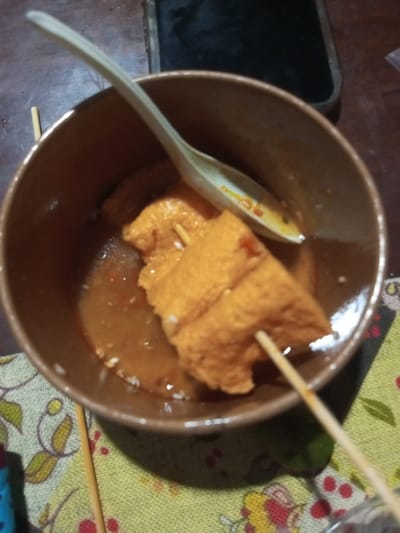 The image displays a close-up, overhead view of a brown ceramic bowl containing a light brown liquid broth with visible red and white particulate matter. Submerged within the broth are at least three irregularly shaped, porous, light orange-brown pieces of food. Two of these pieces are impaled on thin wooden skewers, one protruding upwards from the bowl. A light-colored spoon with faint markings rests in the broth against the far rim of the bowl. The bowl is positioned on a light green fabric surface patterned with yellow and red floral or leaf motifs. In the upper right background, a dark rectangular object resembling a smartphone in a case is partially visible. No individuals are present. The scene is located in Pandaria, India.
