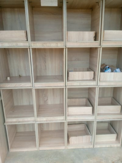 The image shows a close-up, eye-level, full outdoor shot of a large, empty, light-colored wooden shelving unit. The shelving unit is organized into a grid of 12 individual cubbyholes arranged in four rows and three columns. Several of the cubbyholes contain small, empty wooden drawers. The shelves are made of light-colored wood with visible grain patterns. The cubbyholes are open and empty, except for a few drawers and some small cardboard boxes in one of the upper cubbyholes. The background is blurred, but appears to be a concrete or pavement surface. The lighting suggests it is daytime. The overall impression is one of a new, unassembled, or unpacked shelving unit. The location is identified as Indo, Nigeria.