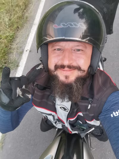 A man wearing a black helmet and a motorcycle jacket rides a motorcycle. The man has a beard and a mustache. He is wearing black gloves and a blue long-sleeved shirt. The motorcycle is gray and black. The man is giving a peace sign with his left hand. The road is gray asphalt with a white line on the left side. There is grass and vegetation on the left side of the road. The scene appears to be outdoors in a rural or suburban area. The image does not provide enough information to determine the specific location in Semypolky, Ukraine, but it depicts a person on a motorcycle on a road.