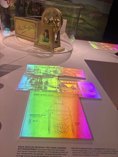Here is a matter-of-fact description of the image:

The image shows a museum display in what appears to be an exhibition related to chocolate or confectionery. A vintage-style machine is presented behind a clear cylindrical enclosure, labeled "Rod Lindt fils". Behind the machine, on the wall, are several illuminated panels. These panels contain text and diagrams. The text is in German and English, with one panel focusing on Nestlé's design for a condenser and describing the process of making sweetened condensed milk. Another panel shows the Nestlé logo and the text "RICHEST II", with a statement about Daniel Peter having a condensing room.

The display is likely located in Kilchberg (ZH), Switzerland, because both Rod Lindt and Nestlé have historical connections to the area. The presence of both names suggests the exhibition focuses on the history of chocolate manufacturing in the region.