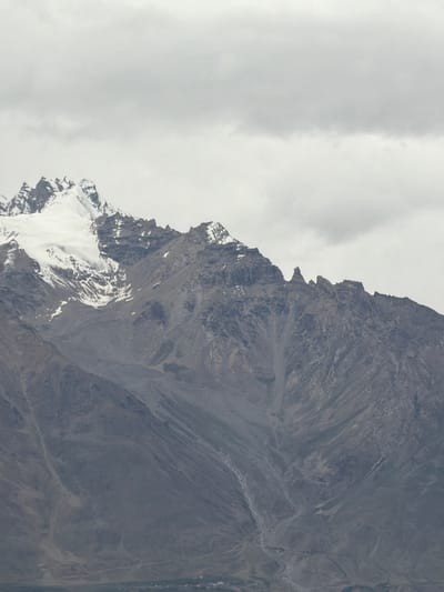 A majestic mountain range under a cloudy sky is captured in this vertical, eye-level shot. The peaks are rugged and dramatic, with snow-capped summits that gleam against the muted gray of the clouds. The mountain slopes are a tapestry of rock and scree, with deep ravines and channels carved into their sides, suggesting a history of erosion and geological activity. In the distance, a small settlement is visible at the base of the mountains, a cluster of buildings suggesting human habitation in this remote landscape. The overall impression is one of vastness, natural power, and the quiet resilience of life in a harsh environment. The location is identified as being in Stongde, India.