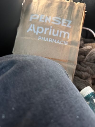 A brown paper bag with white lettering is placed on a person's lap. The bag has the words "PENSEZ Aprium PHARMACIE" printed on it. A person is wearing grey pants, and a blue liquid in a clear plastic bottle with a white cap is visible on the right side of the image. The background is dark and blurred, suggesting an indoor or vehicle environment. The location is Ödsmål, Sweden.