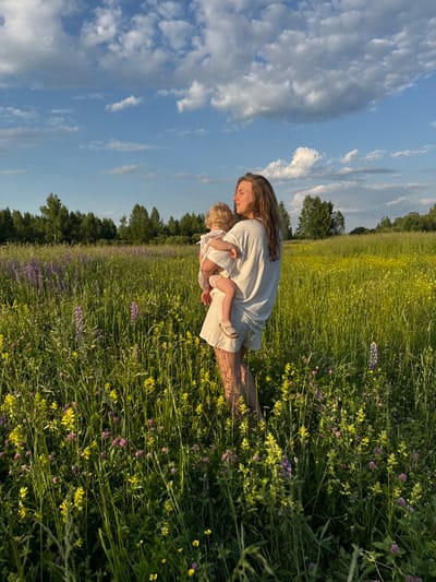 In a sunlit meadow in Koloksha, Russia, a woman with long, wavy brown hair holds a young child. The woman, wearing a loose, off-white outfit and sandals, gazes into the distance, her eyes closed. The child, in a light-colored dress, is held in her arms. The meadow is filled with tall green grass, interspersed with yellow and purple wildflowers. In the background, a line of trees borders the field, and a bright blue sky with scattered white clouds stretches above. The scene captures a moment of quiet connection between mother and child in a natural, rural setting.