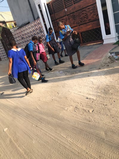 The image captures an outdoor scene, likely a street or pathway, under bright sunlight. The perspective is slightly elevated, looking down along the path.

In the foreground, an adult woman is visible on the right side of the frame, facing slightly left. She is wearing a short-sleeved royal blue top with a white collar and black pants, along with light-colored flat shoes. She is holding a black handbag in her right hand.

To her left and extending further into the background, a line of five young girls, presumably school students, are walking. All of them are dressed in matching school uniforms consisting of a light blue short-sleeved shirt with white collar and details, paired with a grey and white plaid (checkered) skirt. They are all wearing dark socks and black shoes. Each student is carrying a bag; the first student to the left of the adult woman has a colorful, square-shaped lunchbox or small bag. The next student carries a pink backpack, and another further back has a black backpack. The children appear to be walking in a single file, suggesting they are commuting, possibly to or from school, accompanied by the adult.

The immediate background on the left side of the frame features a building. A prominent feature is a large, dark brown wooden gate or door with intricate carved details. Above this gate, there is a white frame or trim. To the left of the gate, a wall with a distinct patterned tile or brick design is visible.

The ground surface dominating the right half and extending towards the foreground is unpaved, appearing to be sandy or dirt. Visible tire tracks and footprints crisscross the dusty surface.

The lighting indicates a sunny day, with clear, somewhat long shadows cast by the individuals, suggesting it might be morning or late afternoon. The overall impression is one of daily life in a residential area, likely in a developing or semi-urban setting, given the unpaved road and style of architecture. No discernible text is visible in the image.