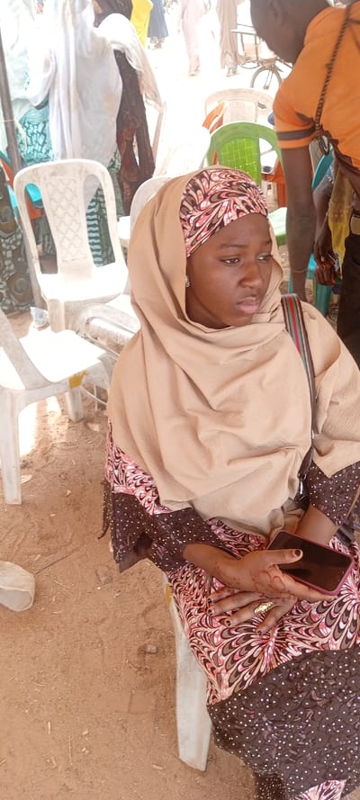 The image captures an outdoor scene in Rijiyar Zaki, Nigeria, featuring a seated young woman in the foreground. She wears a light brown hijab over a patterned underscarf and a dress with a pink, brown, and white patterned upper section, transitioning to a dark brown, white polka-dotted lower half. A beige shawl covers her shoulders. She has small silver-toned stud earrings and a black bag strap with red, green, and brown stripes visible over her right shoulder. Her hands, adorned with henna and dark nail polish, are clasped, with a golden, ornate ring on her right ring finger. She holds a dark-colored smartphone with a reddish case in her right hand. Her gaze is directed towards the viewer's left. The ground is reddish-brown earth. In the background, multiple individuals are visible, many blurred, wearing various garments including white, yellow, and dark fabrics. Numerous plastic chairs, predominantly white, with some green and orange, are scattered throughout the scene. A partial view of a person in an orange t-shirt is seen to the main subject's right. A white plastic container rests on the ground near the left frame edge.