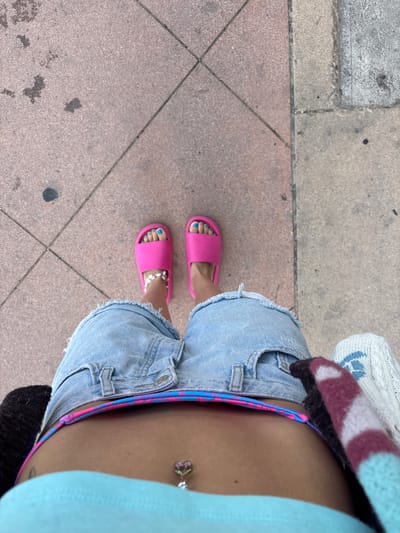 An overhead, first-person perspective image captures a person standing outdoors, likely on a sidewalk or path. The subject's torso, legs, and feet are visible. They are wearing light wash denim cut-off shorts, a light blue top, and a brightly patterned bikini bottom with blue and pink stripes peeking out from the waistband of the shorts. A silver belly button ring, adorned with a pink cherry design and a green stem, is visible on the person's navel.
Below the shorts, the person's feet are clad in bright pink slide sandals. Their toenails are painted a vivid blue. A delicate silver chain anklet is worn on the right ankle. The ground beneath consists of a mix of concrete surfaces, including lighter grey concrete slabs and reddish-brown square tiles with diagonal lines, suggesting a patterned pavement. The overall impression is one of casual summer wear. The lighting indicates it is daytime. The scene is consistent with an outdoor setting in a location like Huntington Beach, United States of America. No specific activity beyond standing is clearly depicted, and no text is visible.