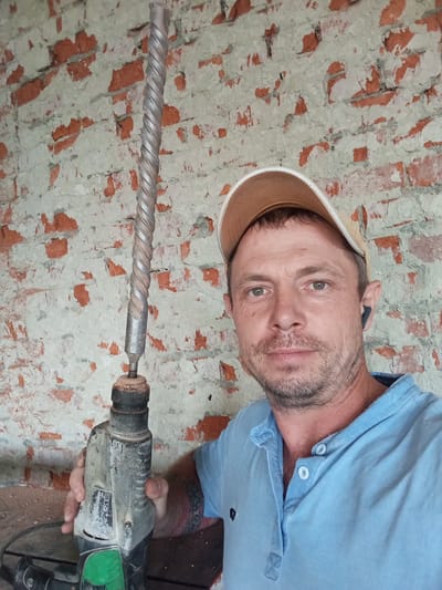 A man is holding a rotary hammer with a long drill bit attached. He is wearing a light blue collared shirt and a tan baseball cap. He also has a black earbud in his right ear. The background is a brick wall that appears to be under construction or renovation. The image was taken in Saratovskaya, Russia.