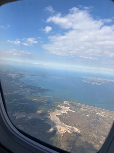The image is an aerial view from an airplane window, looking out over a coastline. The scene depicts the ocean meeting land, with a prominent bay or inlet on the left side of the frame. The land appears to be a mix of vegetation, some dry, barren areas, and bodies of water that form winding channels. In the distance, a more developed coastal area with buildings is visible on a peninsula extending into the sea. The sky above is blue with scattered white clouds. The vantage point suggests the aircraft is flying at a moderate altitude, offering a broad perspective of the landscape and its interaction with the ocean. This specific view is likely of the coastline near Saul, Mozambique, given the characteristic geography of the region with its numerous inlets, bays, and coastal settlements.