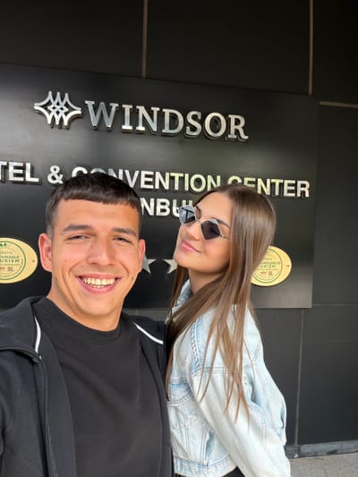 A man and a woman are posing for a photo in front of the Windsor Hotel & Convention Center in Istanbul, Turkey. The man is wearing a black sweater and the woman is wearing a light blue denim jacket and sunglasses. Both are smiling.