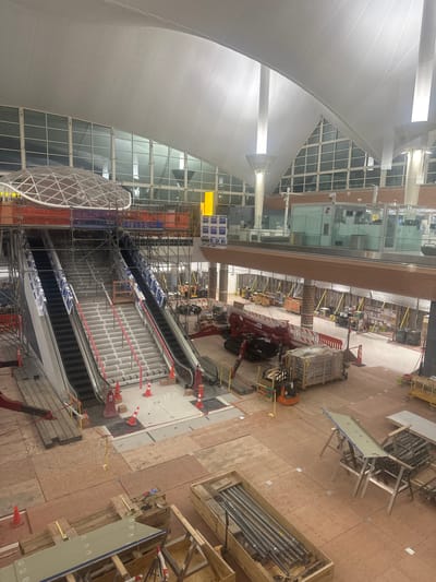 The image shows an overhead view of an interior space under construction. The location is Denver, United States of America. The interior features large windows, a curved roof structure, and multiple escalators under construction. Scaffolding surrounds the escalators. Construction equipment, including a compact crane, is present. Construction materials, such as crates and lumber, are scattered across the floor. There are no visible people. The environment appears to be a large public space, possibly an airport or transit hub undergoing renovations.