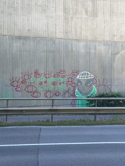 The image depicts a concrete wall alongside a road in Wasserbillig, Luxembourg. The wall is heavily decorated with graffiti, including a large smiling face, a cluster of eye-like shapes, and some text in green. A metal guardrail runs along the edge of the road, separating it from the wall. Beyond the guardrail is a paved road with a solid white lane divider. Patches of grass and vegetation grow between the road and the guardrail. The concrete wall consists of two different sections with different textures.