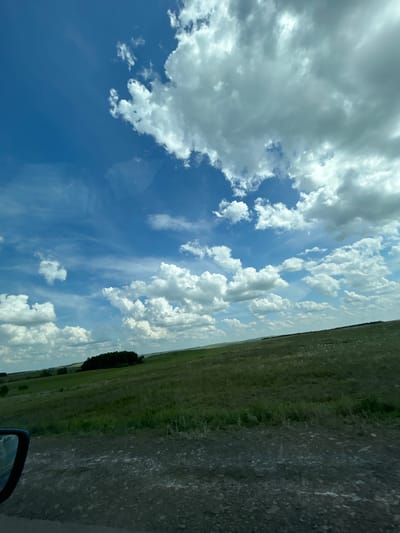 The image depicts a wide, rolling landscape under a bright blue sky filled with scattered cumulus clouds. The foreground is a rough, gravelly dirt road, with the dark housing of a side mirror visible on the left. Beyond the road, a lush green grassy field stretches into the distance, with gentle slopes and undulations. A cluster of dark green trees is situated on a further hill. The horizon is a soft line where the green fields meet the expansive sky. There are no people visible in the image. The scene appears to be a rural or natural environment, likely in the city of Ishmurzino, Russia.