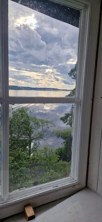 This image shows a view through an old, multi-paned window, looking out onto a natural landscape. The window frame is painted white and appears worn, with some visible cracks in the wooden frame and along the wall above it. On the right side of the frame, a small, block-shaped piece of wood is attached to the wall.

The scene outside the window is dominated by reflections on the glass, creating a layered effect. The left pane primarily shows a reflection of a cloudy sky with dramatic light breaking through, suggesting a time around sunrise or sunset. This reflection also mirrors elements of the landscape.

The right pane reveals a body of water, likely a lake or river, reflecting the sky and the surrounding trees. Lush green foliage, including the branches of a large tree, is visible in the foreground and middle ground. The water's edge appears to be lined with reeds or tall grasses. In the distance, a strip of land with more trees and possibly buildings can be seen across the water.

The overall impression is one of a serene, natural setting viewed from an indoor, perhaps rustic, location. The interplay of reflection and reality creates a visually interesting and slightly ethereal quality to the scene. The weather appears to be mixed, with dramatic clouds and sunshine. The location context of Sigtuna, Sweden, suggests a possible setting in a rural or waterside area of this historic town. There is no visible text in the image.