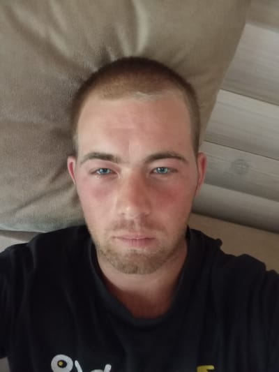 A close-up, overhead selfie of a young Caucasian man is shown. He has fair skin, short brown hair, and blue eyes, and he is wearing a black t-shirt. His face is slightly flushed, and there is a faint stubble on his chin. The man is lying on a textured surface that appears to be a bed or couch, with a wooden-slatted surface visible in the background. The image was taken in Sokyrna, Ukraine.