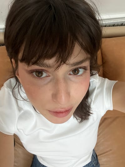 A close-up, high-angle portrait captures a single individual in what appears to be an indoor setting in Le Pré-Saint-Gervais, France. The subject is a young adult female with dark brown, shoulder-length hair styled with bangs covering the forehead. Her eyes are brown and gaze directly upward into the camera. She exhibits light skin with visible freckles across her cheeks and nose, and wears light makeup including what appears to be blush and lip gloss. She is dressed in a white, short-sleeved crew-neck t-shirt, with blue denim fabric visible at the bottom of the frame. The background consists primarily of a light brown or tan upholstered surface, likely a sofa or cushion, to the subject's right, and a sliver of a light-colored wall or radiator at the top edge. The lighting is even, suggesting ambient indoor illumination. No other persons or specific actions beyond posing for the photograph are evident.